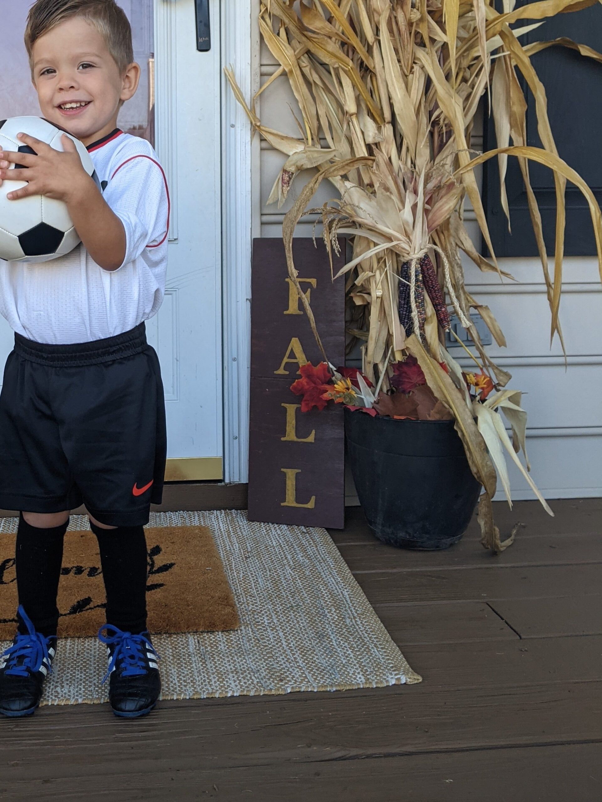 young boy holding a soccer ball in front of  fall decorations that are corn stalks