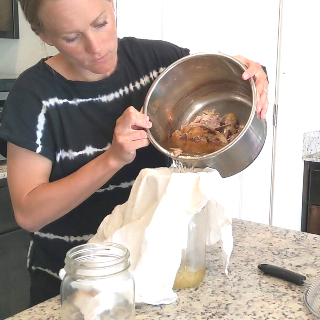 woman wearing a black shirt pouring chicken bone broth into a tea towel funnel into a large mouth mason jar in her kitchen