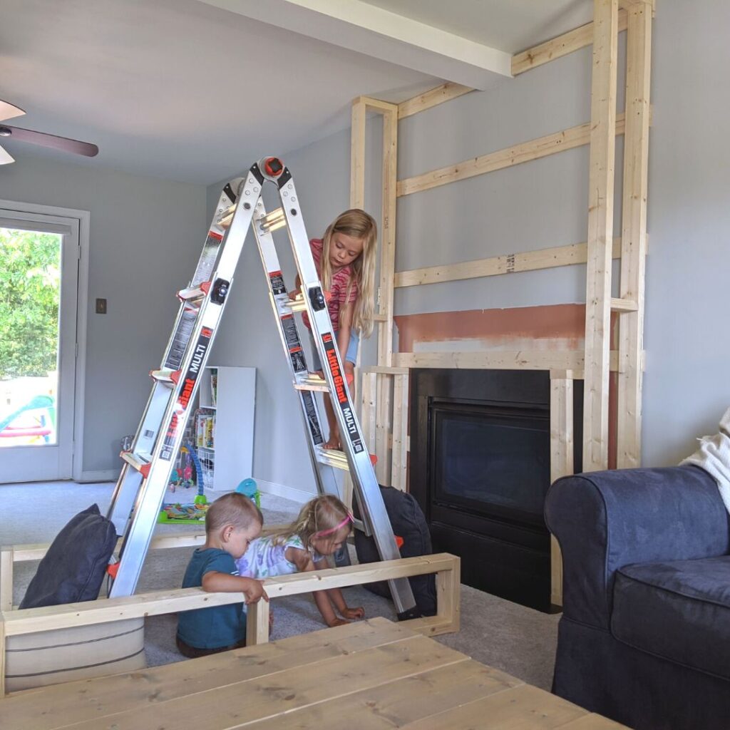 kids standing on a little giant ladder near a fireplace makeover frame in progress with a blue couch next to them.