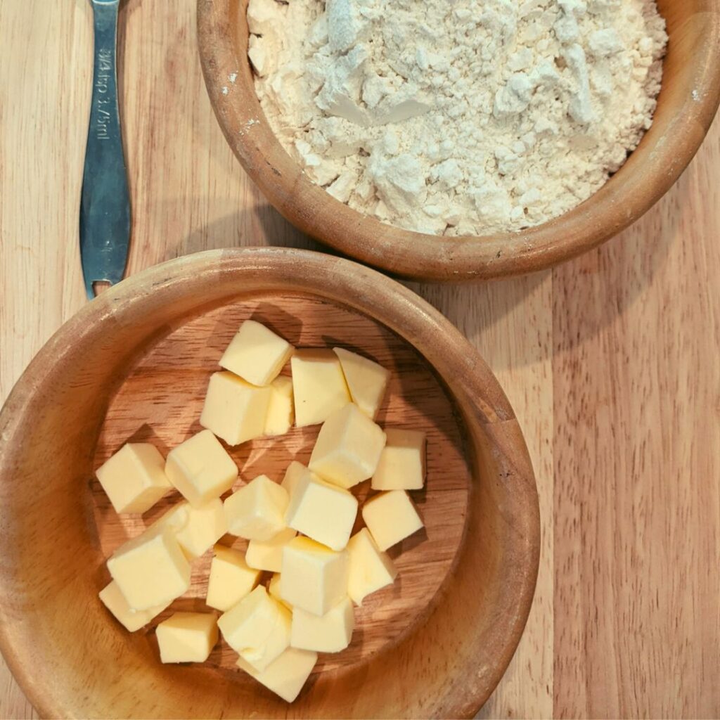 cubed butter in a wooden bowl. flour in a wooden bowl both on a wooden cutting board