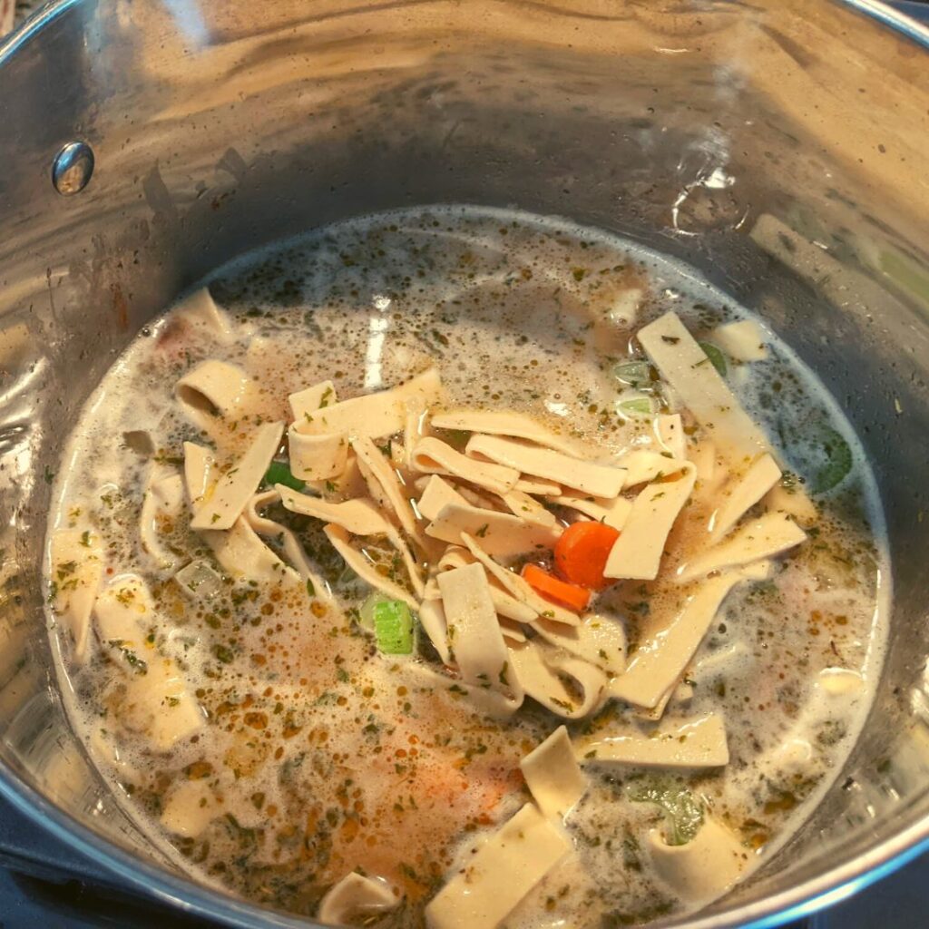 chicken noodle soup with wide egg noodles, celery, carrots, onions, fresh parsley, sage, salt, black pepper inside a large stock pot.