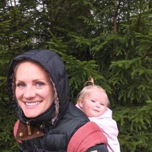 woman standing in front of a real christmas tree while holding her child in an ergo carrier