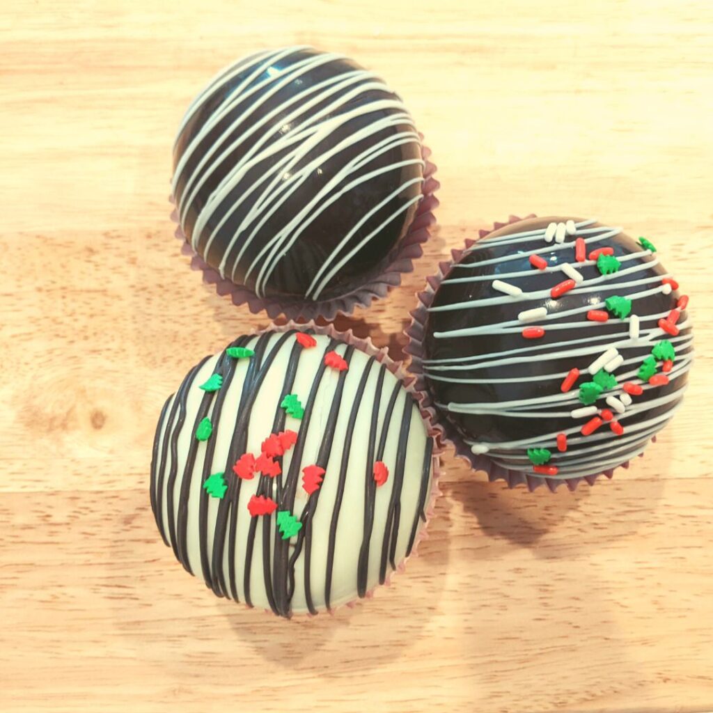 Three hot chocolate bombs in cup cake liners with christmas sprinkles on top sitting on a wooden cutting board