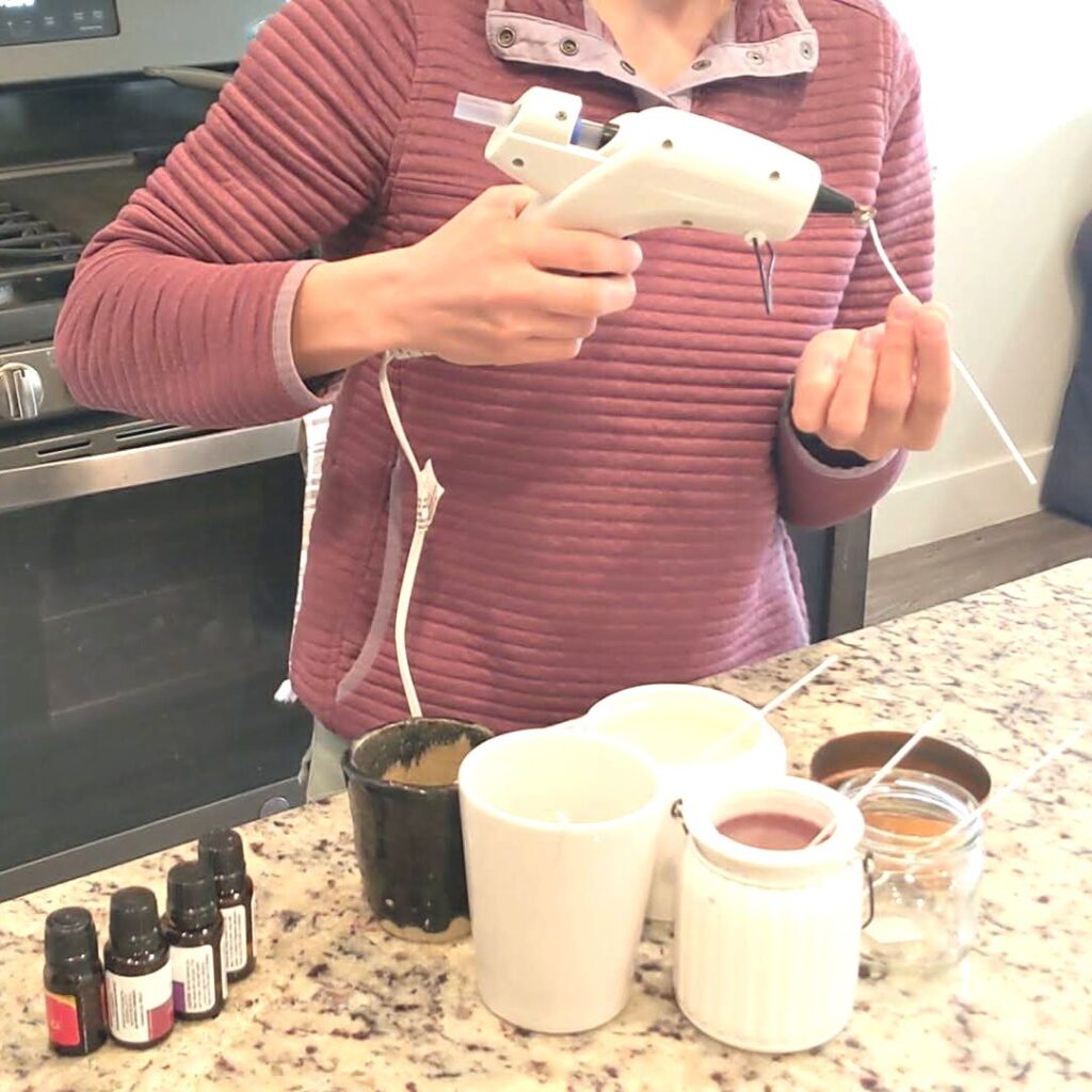woman using a glue gun to put glue on the bottom of the wick that is in her hand before she puts it into her candle jars that are on the counter top next to her essential oils